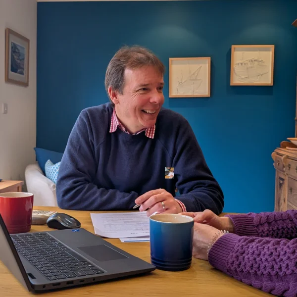 Chartered Financial Adviser Kris Booth is smiling as he chats to a client who is not visible. A laptop and mugs can be seen on the table.