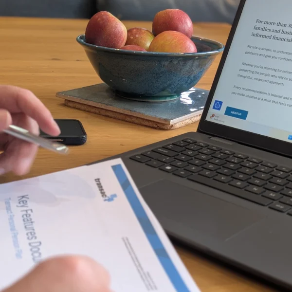 A laptop is open on a table, with a bowl of apples behind it. In the foreground a sheet of life insurance information, is visible, with a pair of hands showing too.