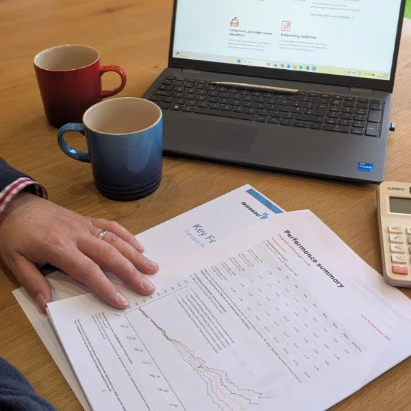 A laptop is open on a table, with a red mug and a blue mug. In the foreground are sheets of information about savings, investments and ISAs, with one hand and a calculator showing too.